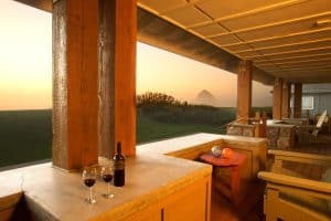 Covered coastal patio with wine glasses, wooden chairs, sunset light, and ocean view in the distance.