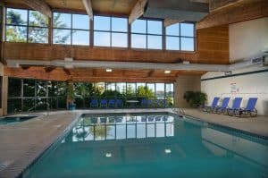 Indoor hotel pool with large windows, wood ceiling beams, lounge chairs, and natural light.