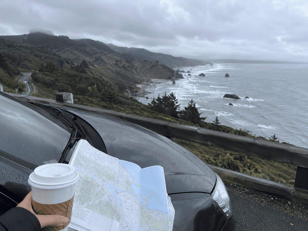 A hand holds a coffee cup and an open paper road map on a car hood at a foggy Oregon Coast overlook, with Highway 101 winding along the shore and sea stacks below
