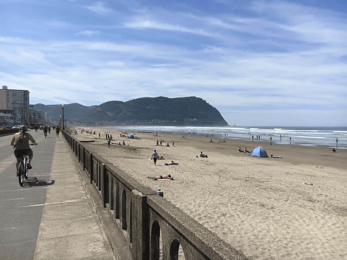 A cyclist rides the Seaside Promenade beside the sandy beach with Tillamook Head in the distance on a sunny summer day
