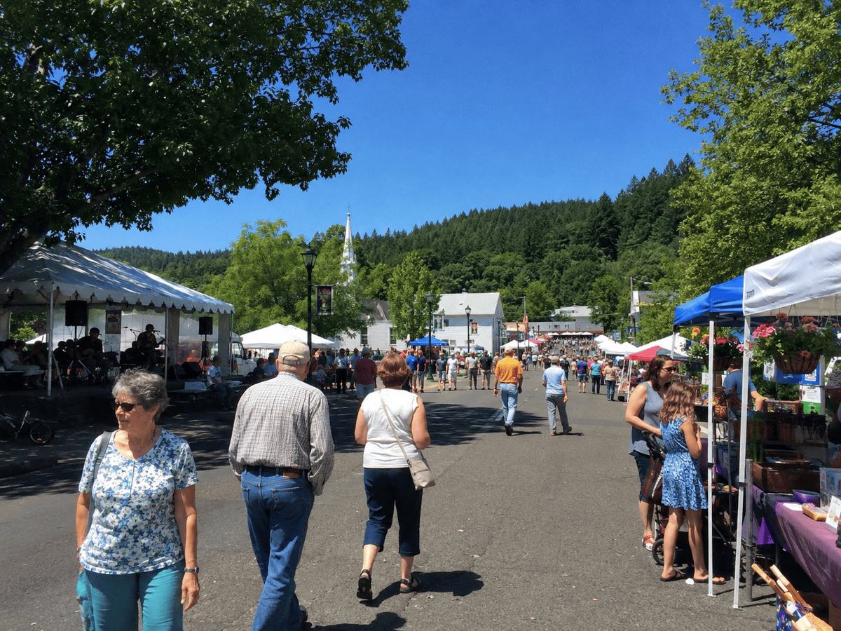 Crowds walk a tree-lined main street with vendor tents and live music at the Toledo Summer Festival, a church steeple and forested hills behind