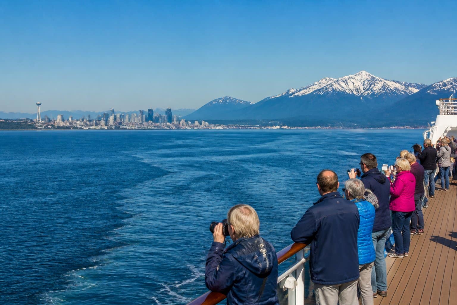 A large cruise ship sailing on calm blue waters with snow-covered mountains and the Seattle skyline in the background.