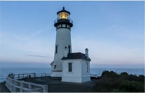 The tall white Yaquina Head Lighthouse near Newport, Oregon, glowing at dusk above the Pacific Ocean