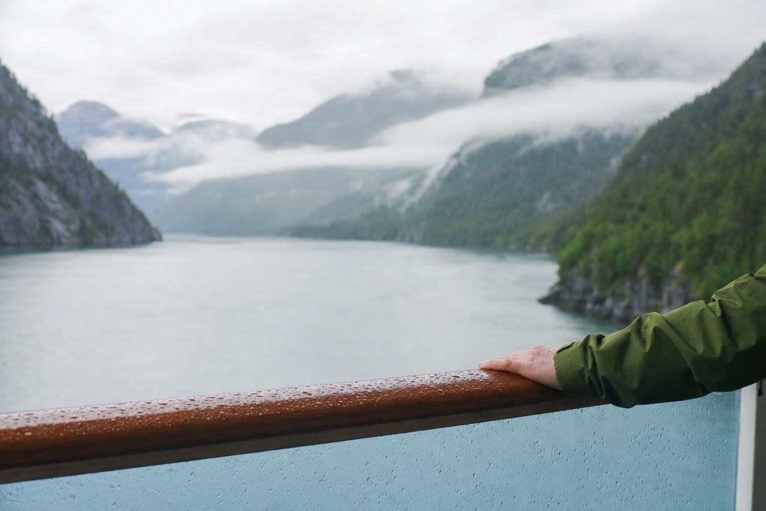 Misty Alaska fjord seen from a cruise balcony with a rain-jacket sleeve on a wet railing