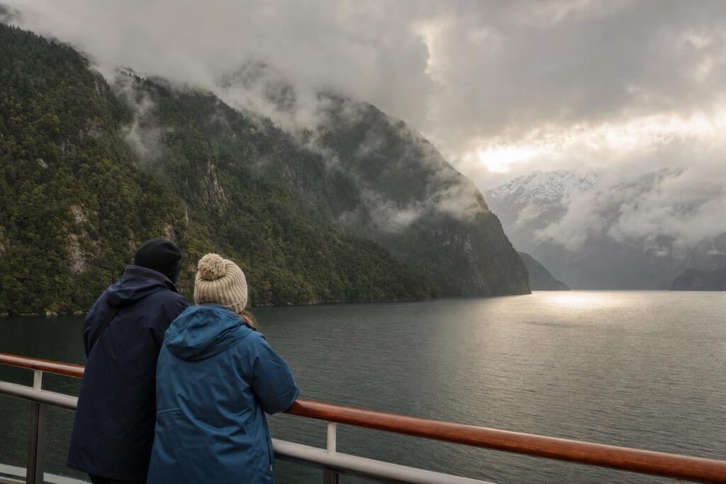 Passengers in rain jackets on a ship deck looking at a misty Alaska fjord with forested mountains