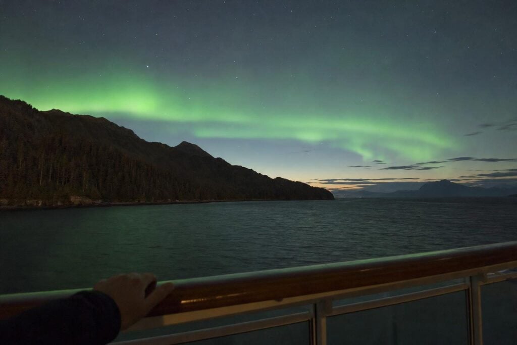 Green northern lights glowing over dark water and mountains seen from a balcony on an Alaska cruise