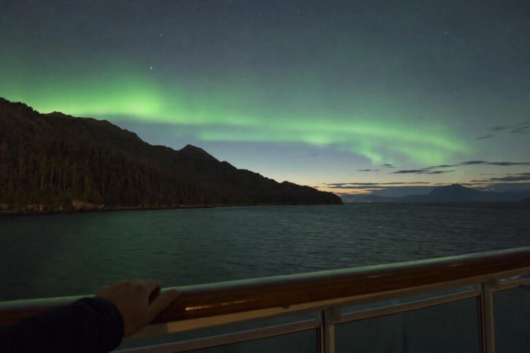 Green northern lights glowing over dark water and mountains seen from a balcony on an Alaska cruise