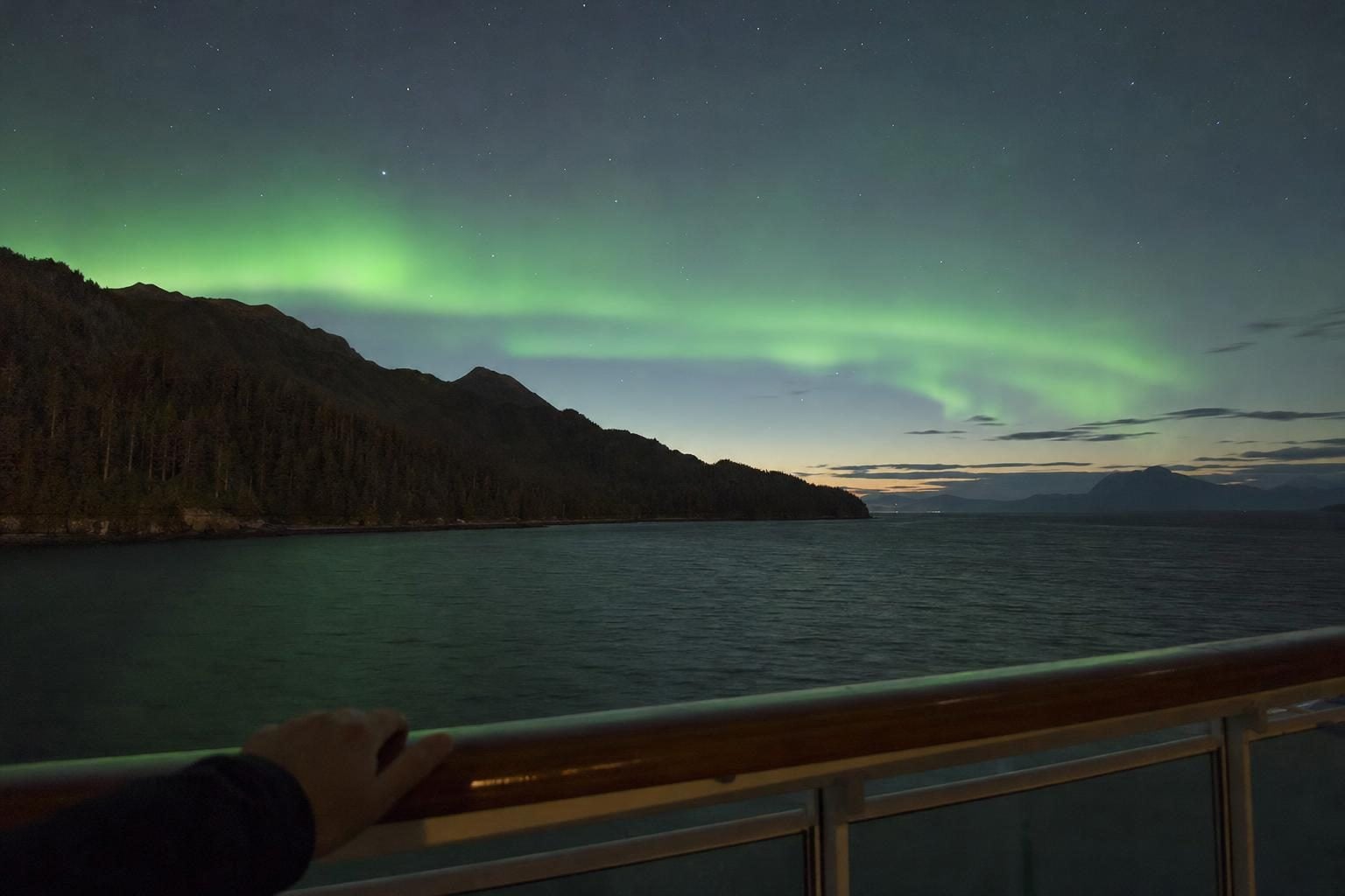 Green northern lights glowing over dark water and mountains seen from a balcony on an Alaska cruise