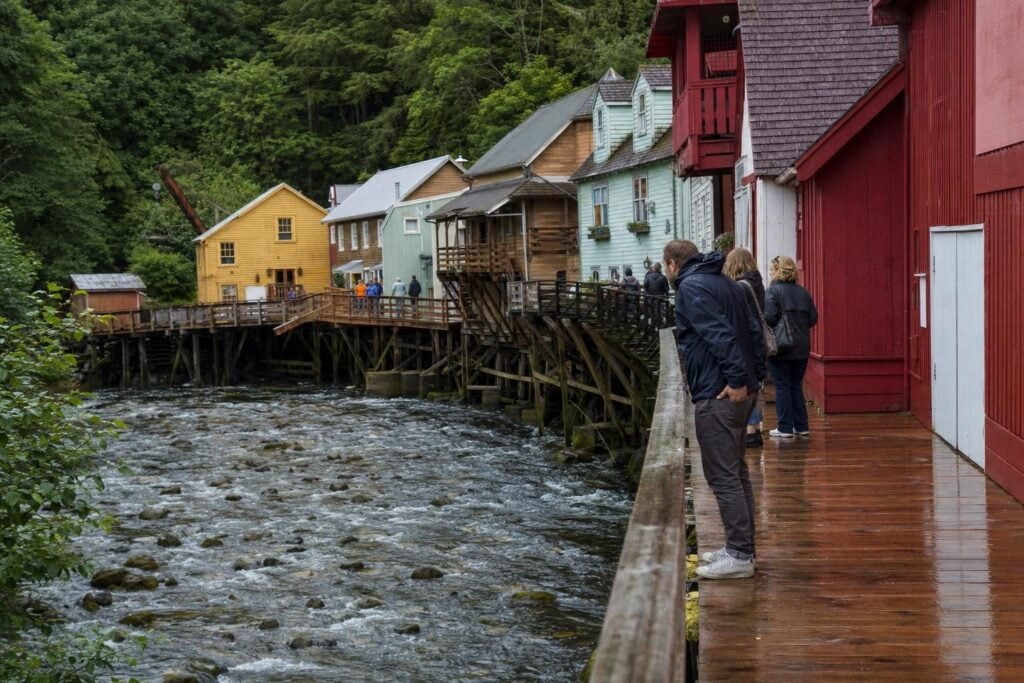 Visitors in rain jackets on a wooden creekside boardwalk with historic painted buildings in an Alaska cruise port