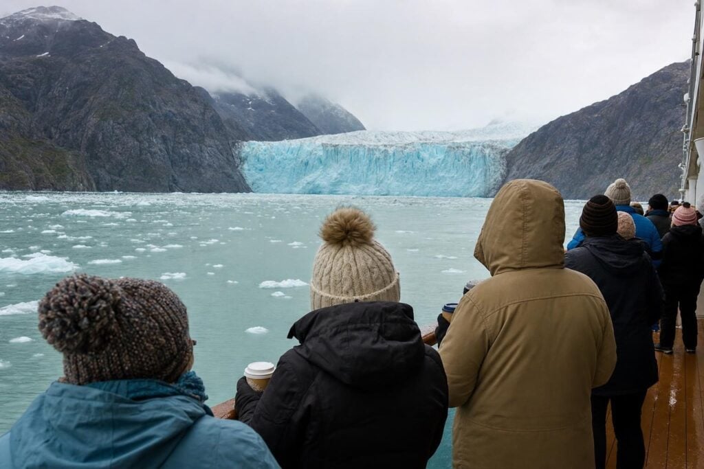 Passengers in warm jackets on a ship deck watching a tidewater glacier during an Alaska Inside Passage cruise