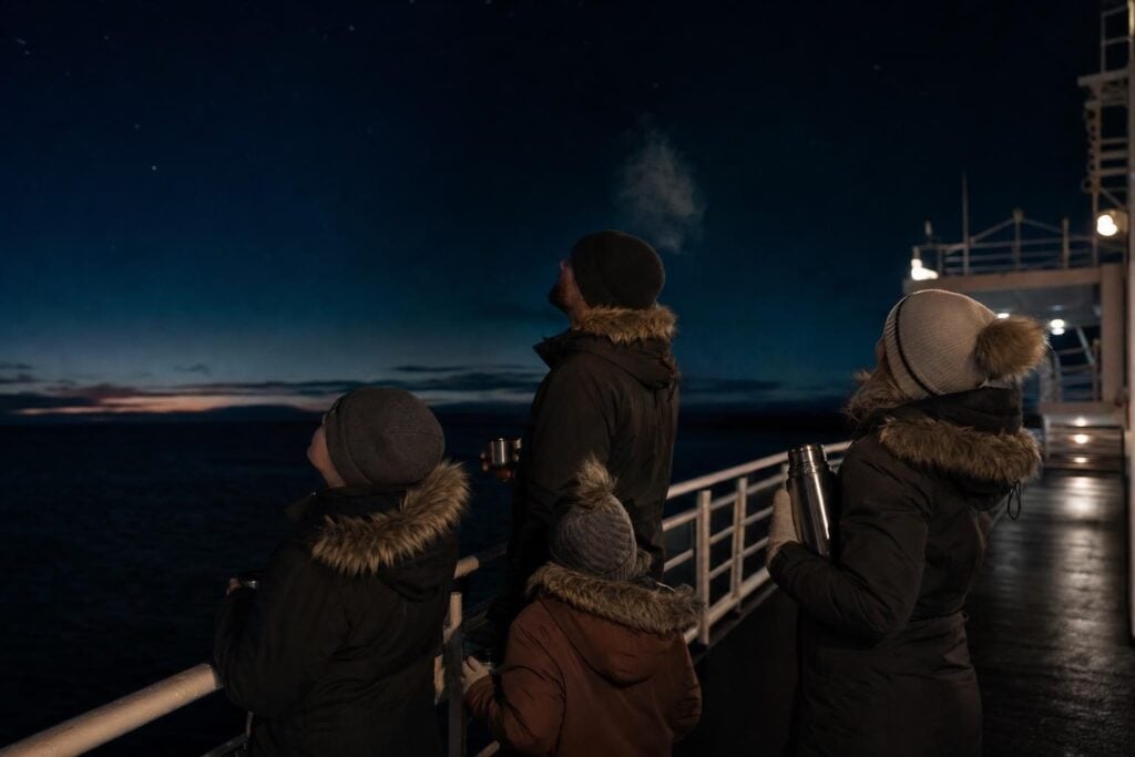 Family in warm jackets and beanies on a dark cruise ship deck watching the night sky in Alaska