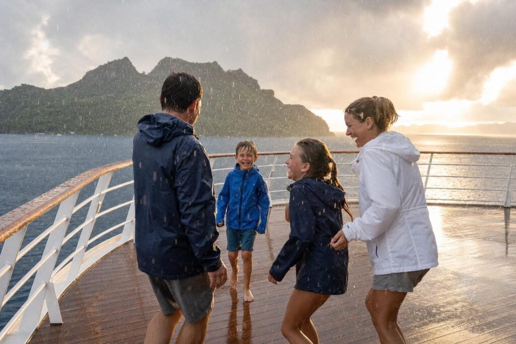 Family in light rain jackets on a cruise ship deck during a passing tropical shower near a green island