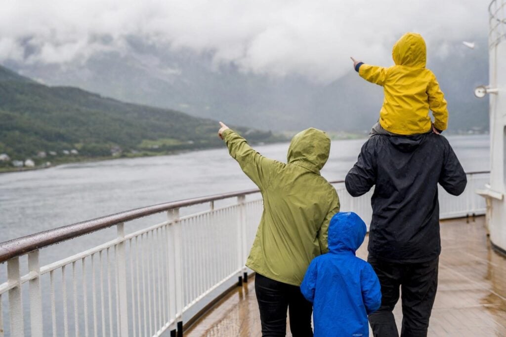 Family in rain jackets on a cruise ship deck watching an Alaska shoreline in light rain