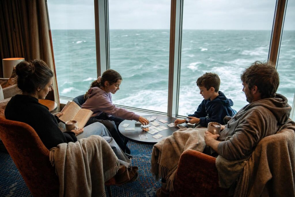 Family playing cards by a lounge window on a sea day during a longer West Coast Alaska cruise