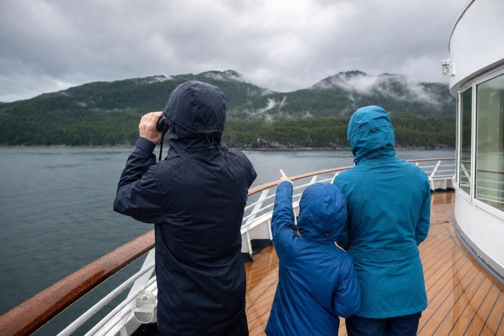 Family in rain jackets using binoculars on a cruise ship deck to scan an Alaska shoreline