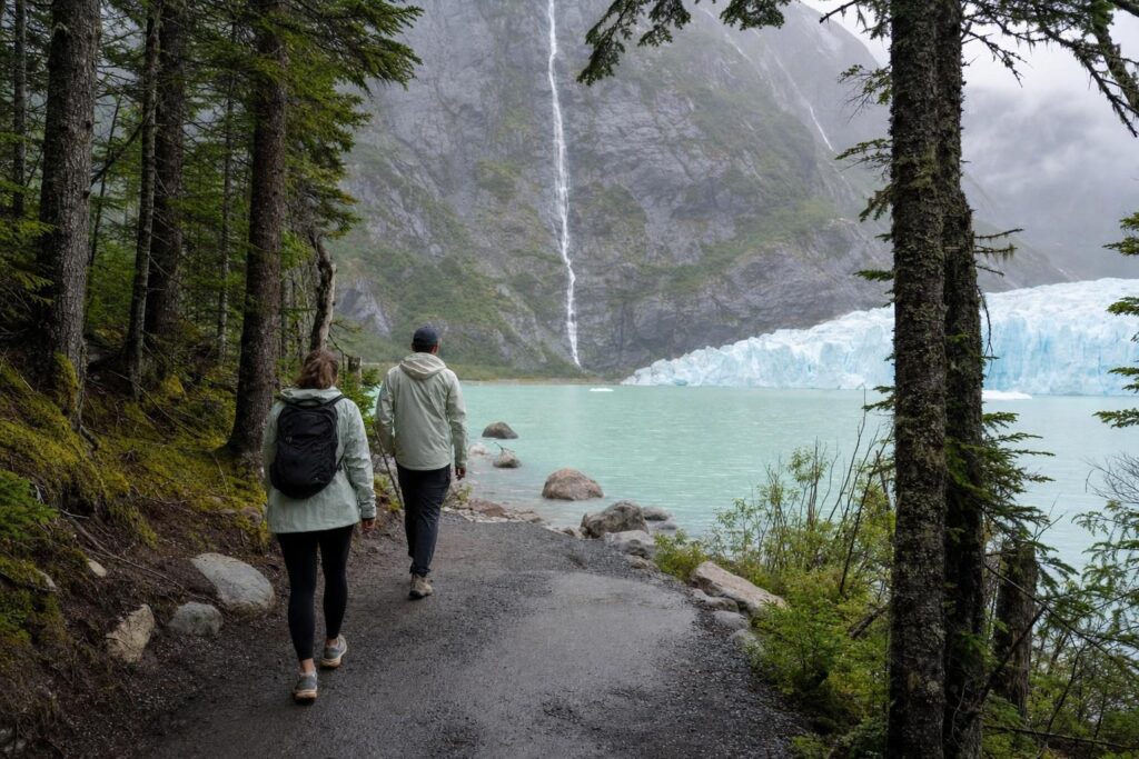Two travelers hiking a forest trail toward a glacier lake and a tall waterfall
