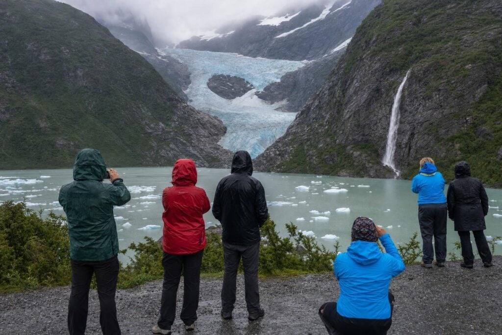 Visitors in rain jackets at a glacier viewpoint with a waterfall and icebergs on a glacial lake