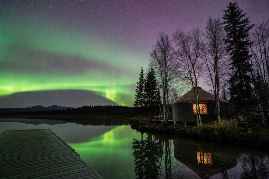 Green aurora over a calm lake and a glowing yurt in interior Alaska, the best place to see northern lights
