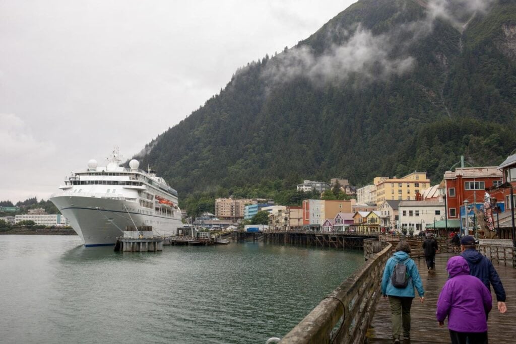 Large white cruise ship docked beside a colorful historic downtown waterfront with a forested mountain behind