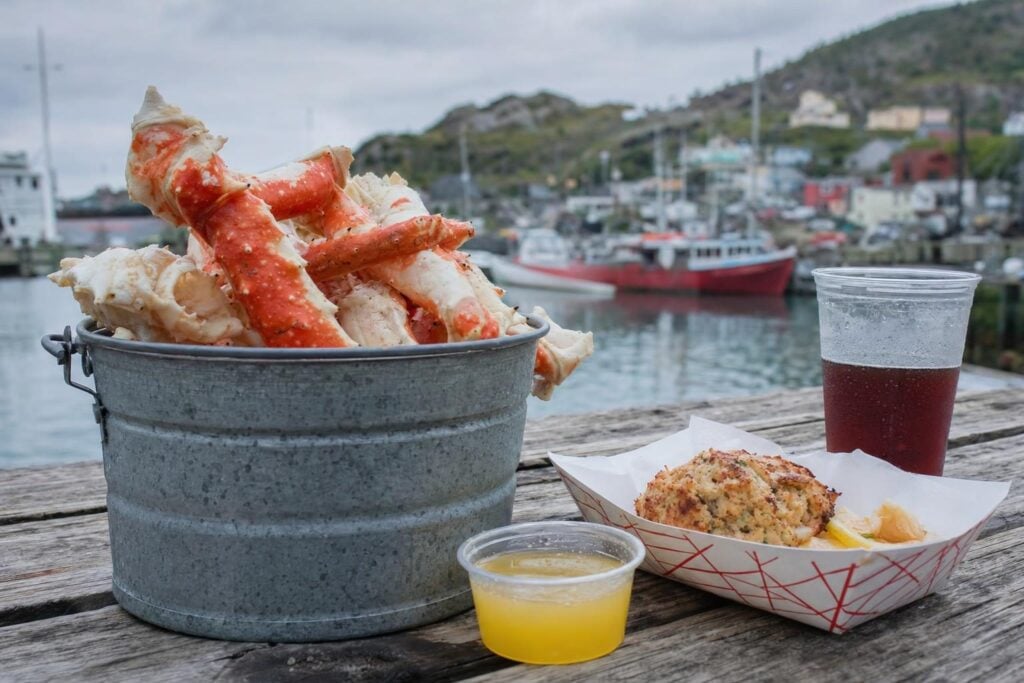 Steamed king crab legs, melted butter and crab cakes on a wooden table beside a working harbor