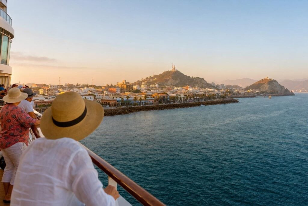 Cruise ship approaching a Mexican Pacific port city at sunrise with a waterfront and lighthouse on the hill