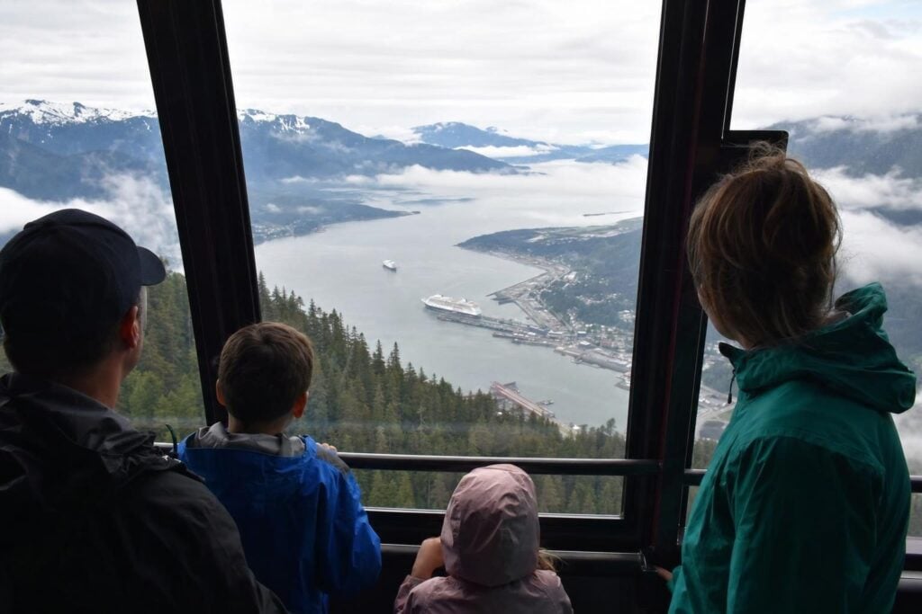 View from an aerial tram cabin over a harbor with a cruise ship far below and snowy peaks in the distance