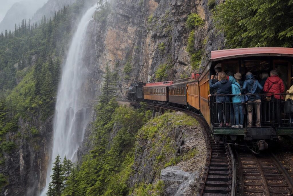 Vintage narrow-gauge excursion train curving along a gorge ledge beside a misty waterfall