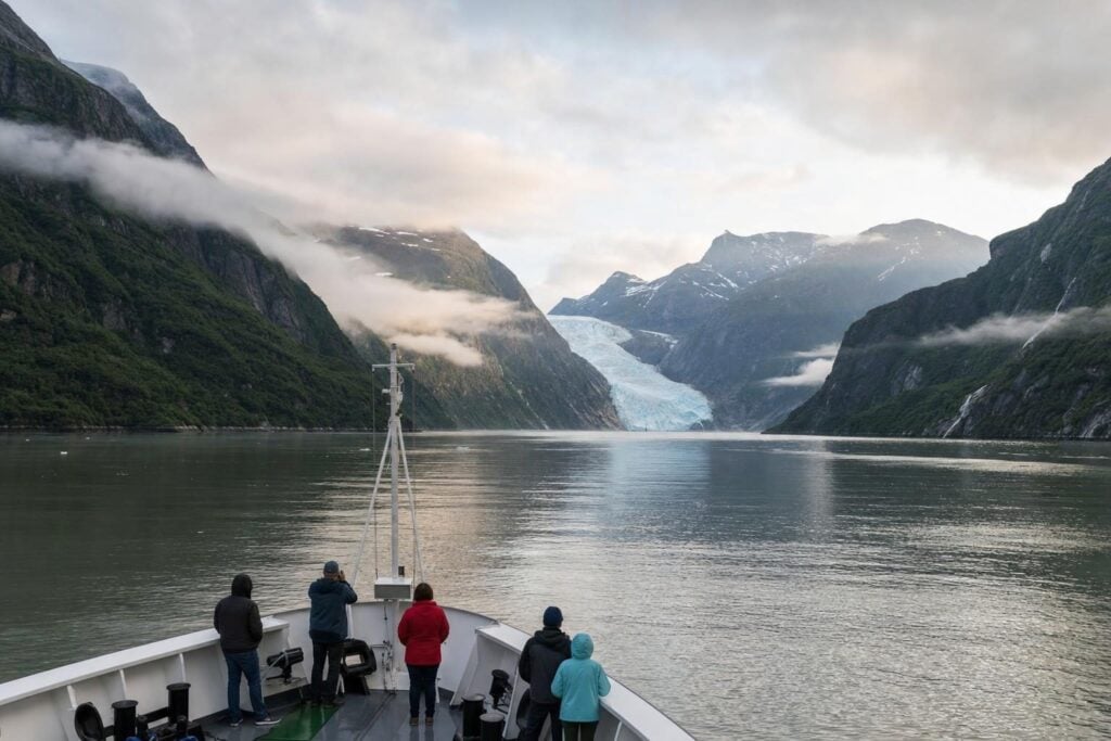 Small ship Alaska cruise gliding through a misty fjord with forested mountains and a distant glacier