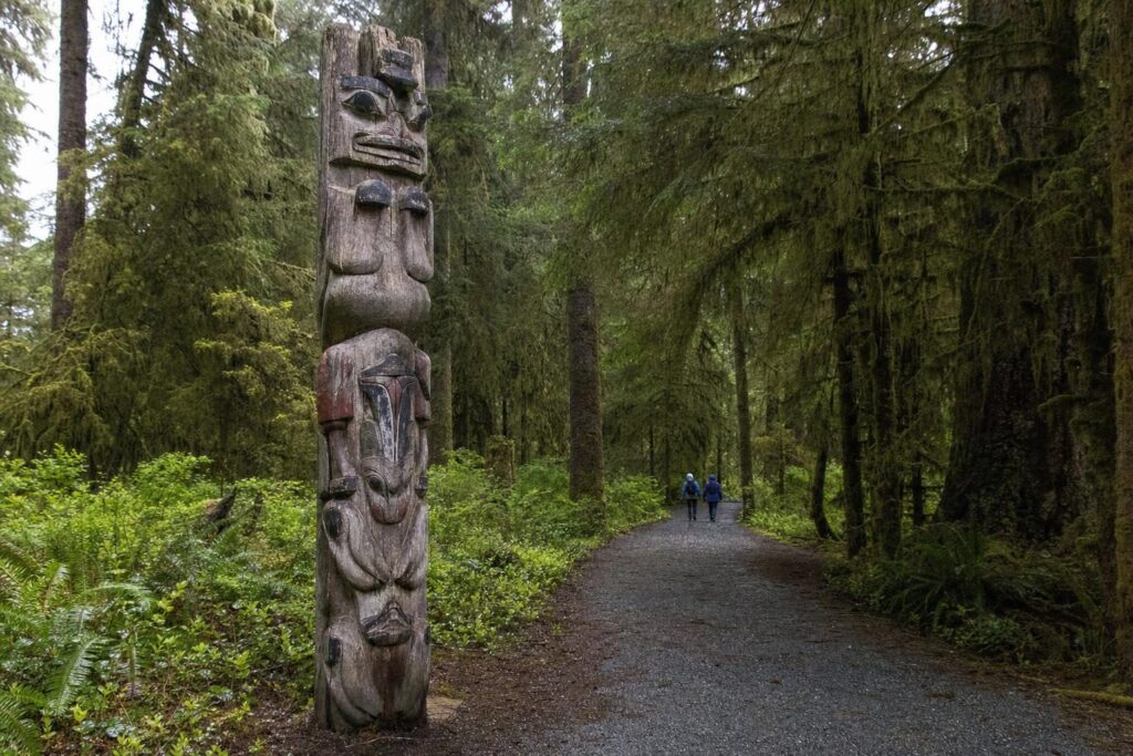 Carved totem pole beside a mossy rainforest trail with two walkers in rain jackets in Southeast Alaska